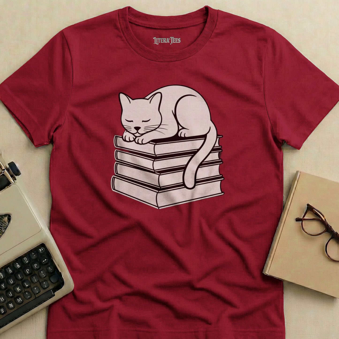 White cat sleeping on a stack of books, printed on a maroon t-shirt.