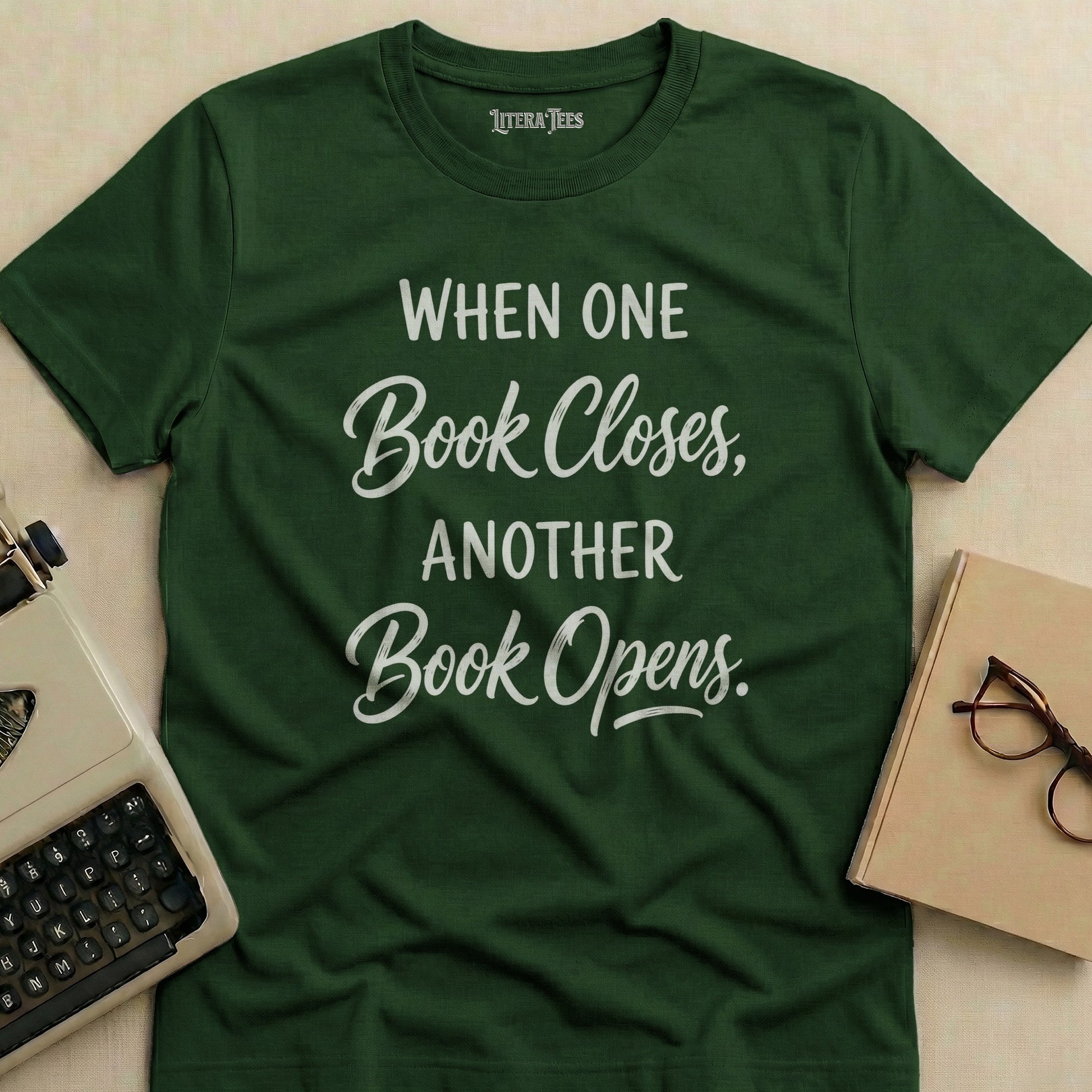 Dark green t-shirt with white text "When One Book Closes, Another Book Opens."