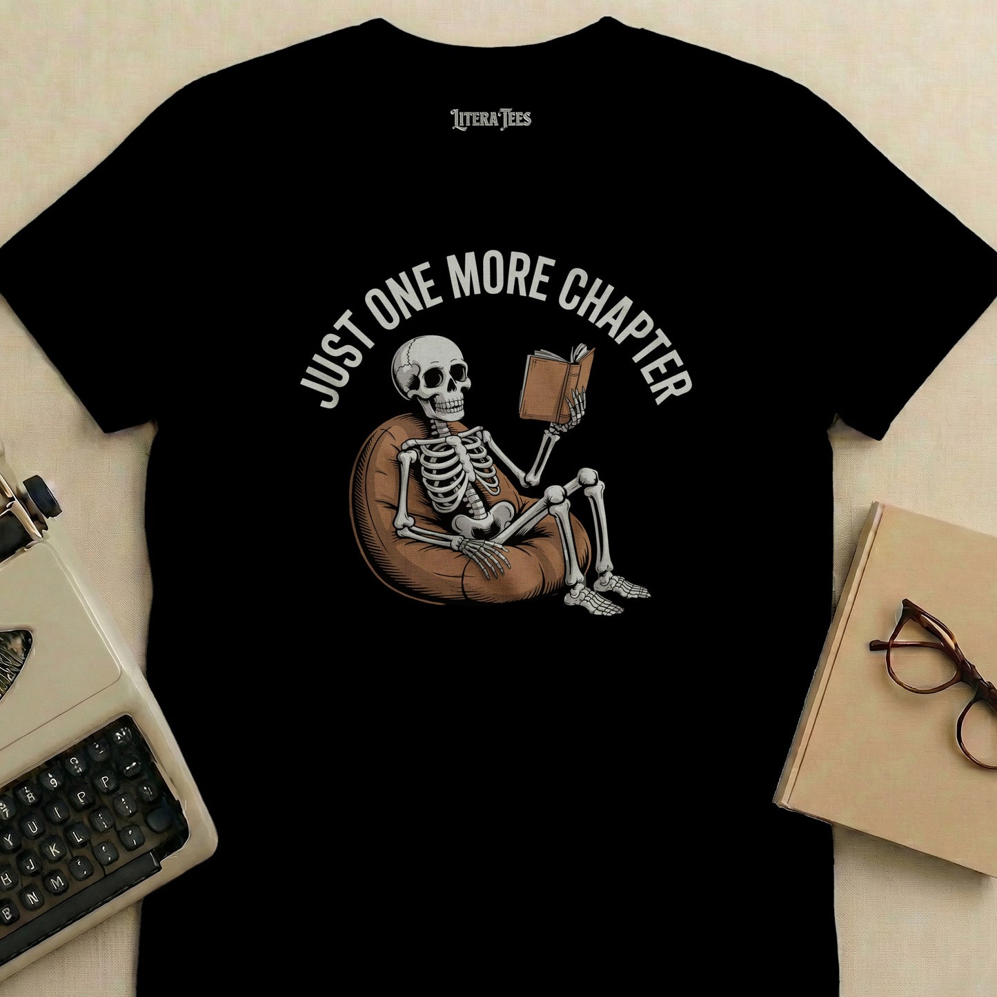 Black t-shirt with skeleton reading on beanbag, "Just One More Chapter" text.