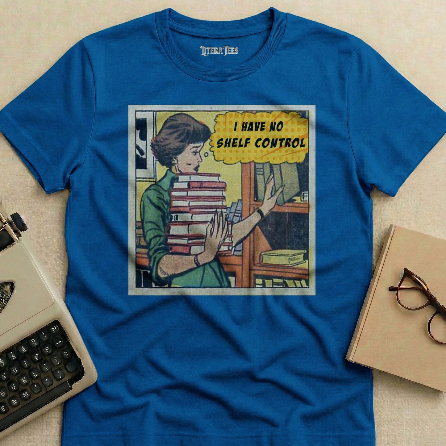 Blue t-shirt with a comic-style graphic of a woman holding books and the text "I HAVE NO SHELF CONTROL".
