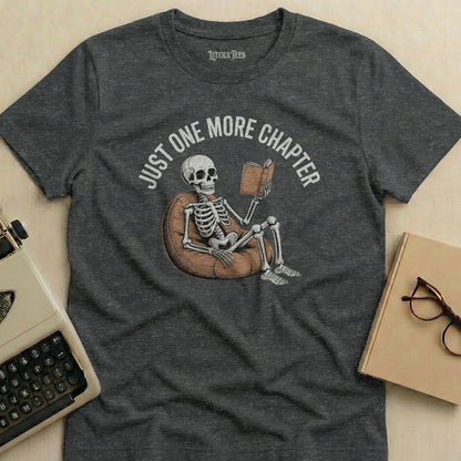 Dark grey t-shirt with skeleton reading a book, text "JUST ONE MORE CHAPTER".