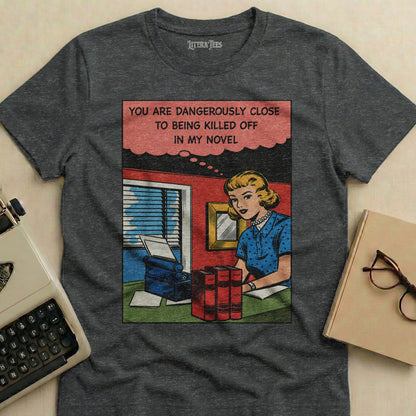 Dark t-shirt with comic panel of woman at desk, saying "YOU ARE DANGEROUSLY CLOSE TO BEING KILLED OFF IN MY NOVEL".