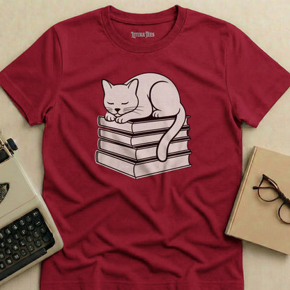 White cat sleeping on a stack of books, printed on a maroon t-shirt.