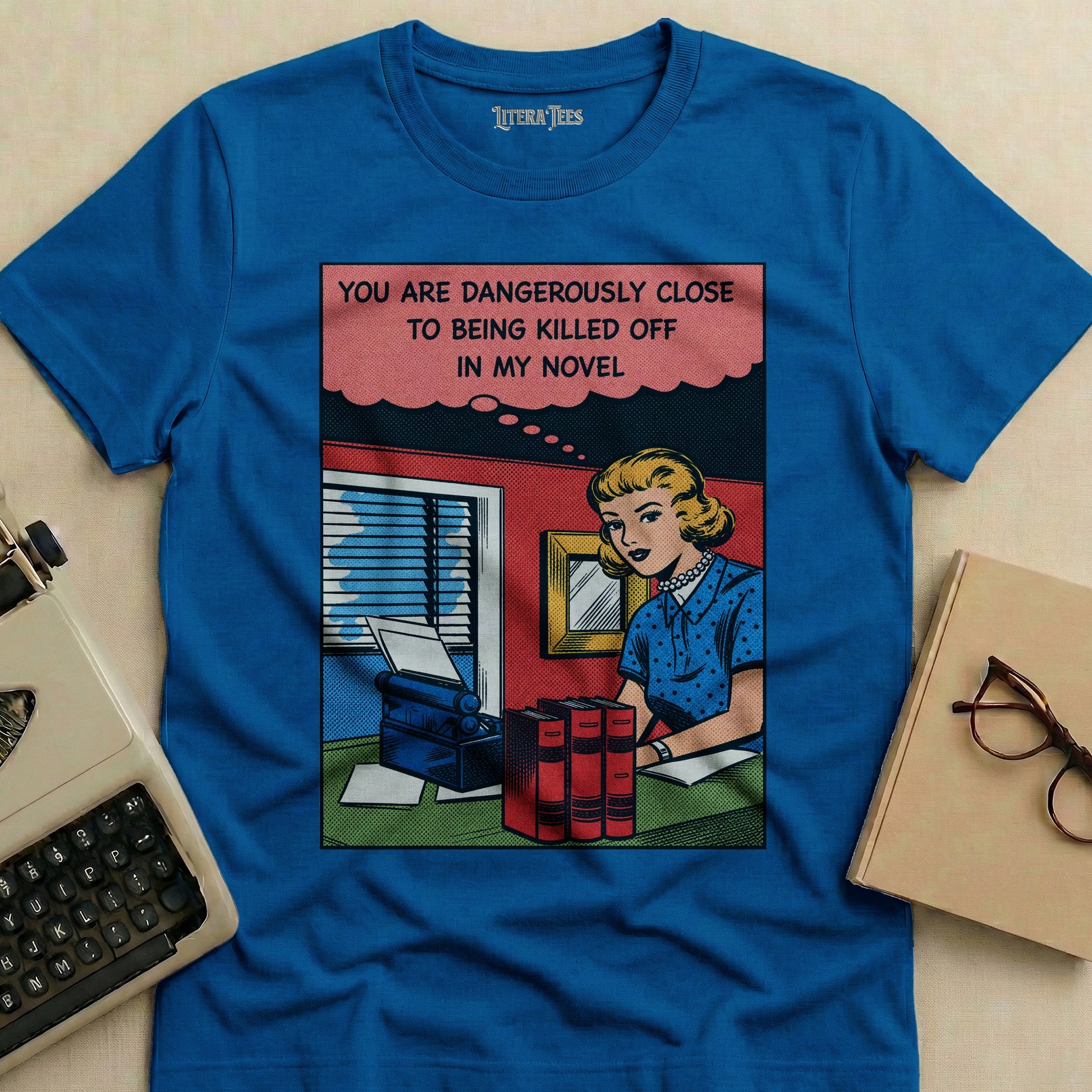 Blue t-shirt with comic art of a woman at a desk, reading "YOU ARE DANGEROUSLY CLOSE TO BEING KILLED OFF IN MY NOVEL."