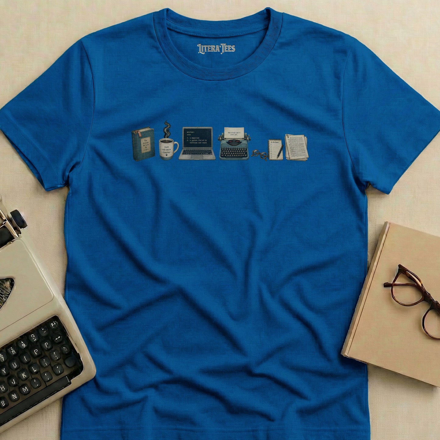 Blue t-shirt with writer-themed illustration of books, coffee, laptop, typewriter, and glasses.