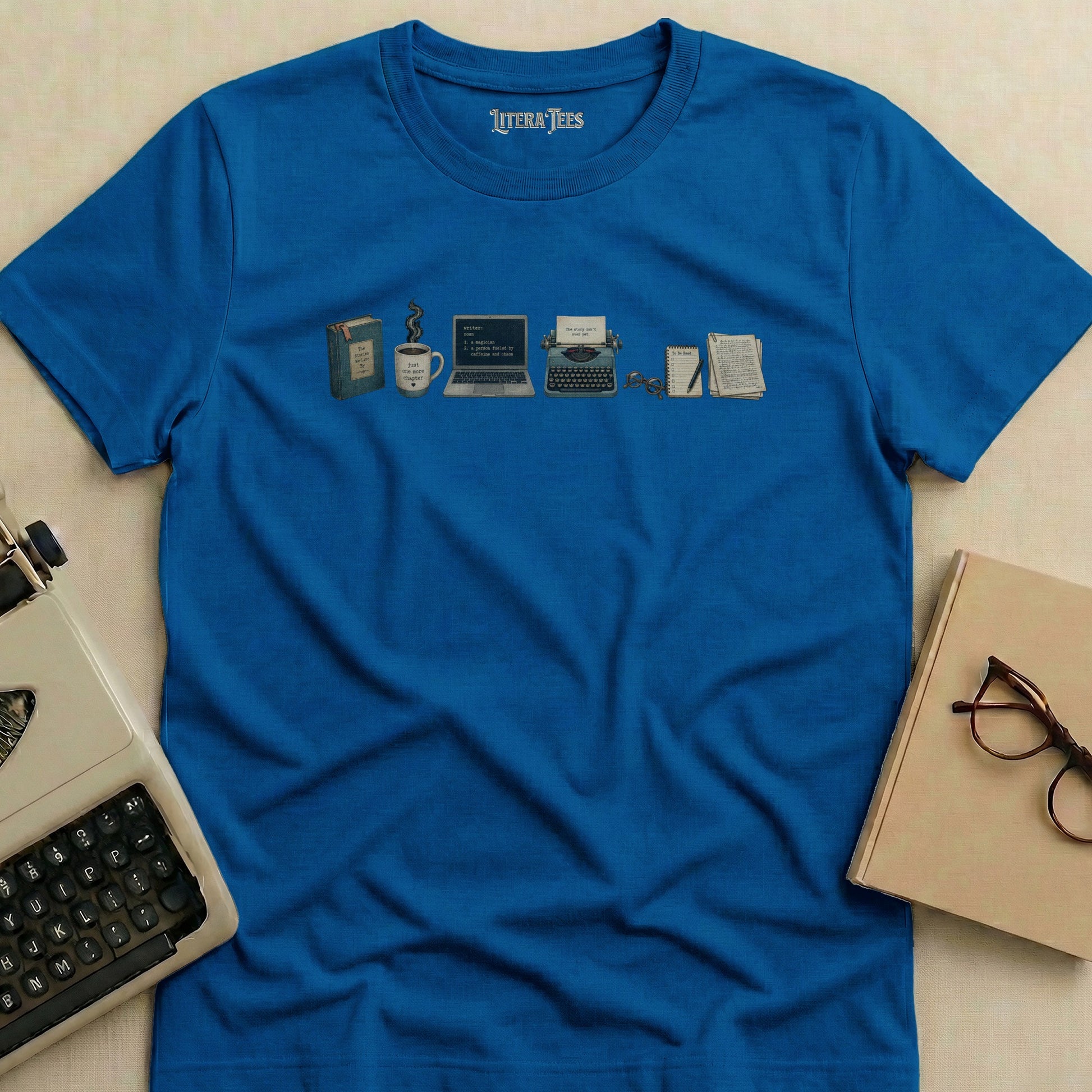 Blue t-shirt with writer-themed illustration of books, coffee, laptop, typewriter, and glasses.