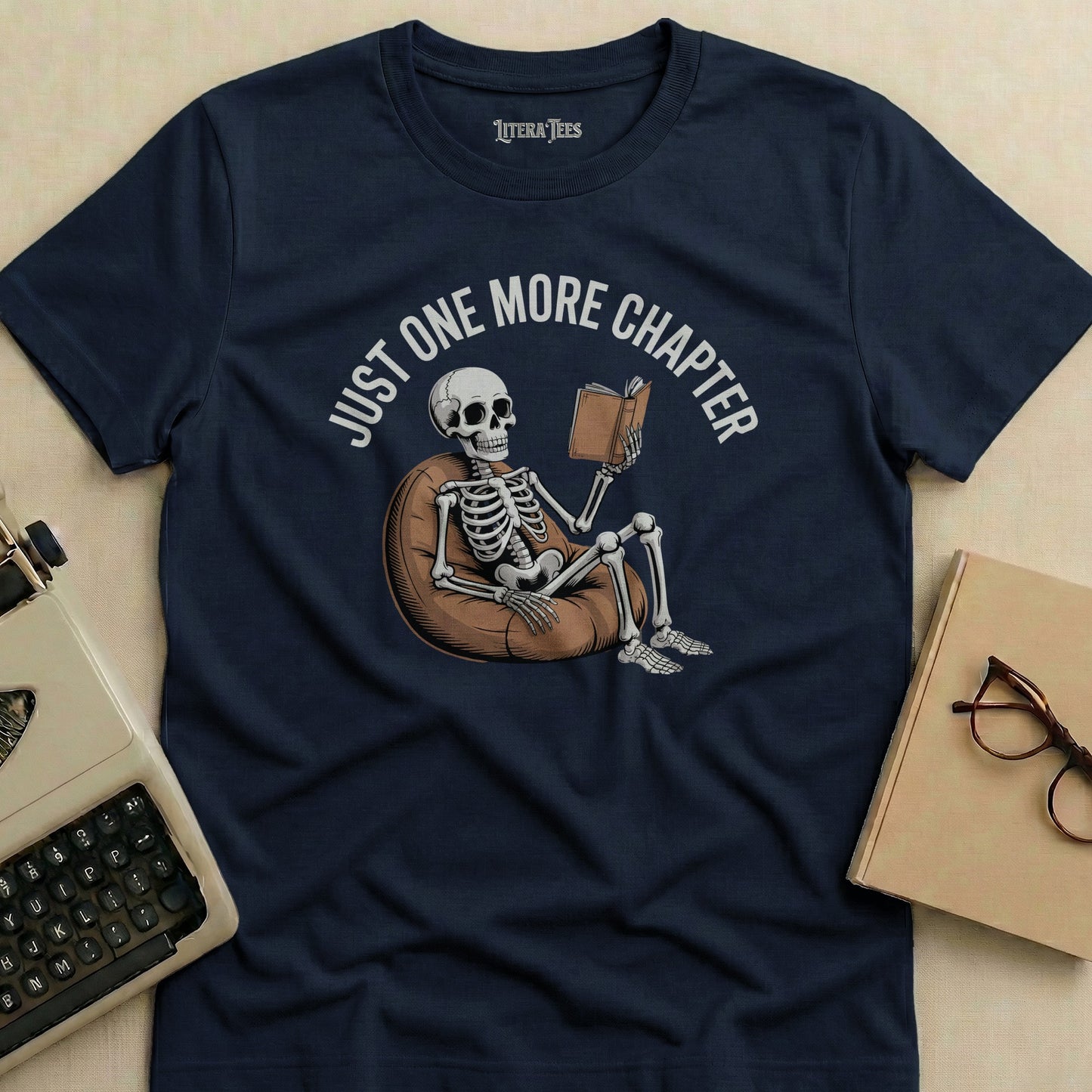 Dark blue t-shirt with "JUST ONE MORE CHAPTER" and skeleton reading on beanbag.